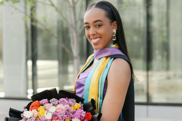 A woman in a graduation gown proudly holds a bouquet of flowers, celebrating her academic achievement.