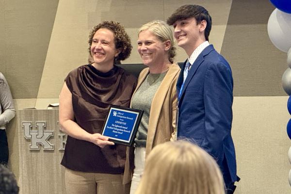  UK's Acting Vice President for Research Ilhem Messaoudi (left) and student nominator Brayden Nichols (right) present Lindsey Fay (center) with a commemorative plaque recognizing her as a 2025 Undergraduate Mentor of the Year..