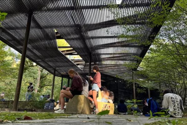 People gather beneath a shaded metal pergola in a leafy park, sitting on rocks and benches with backpacks, creating a relaxed outdoor mood.