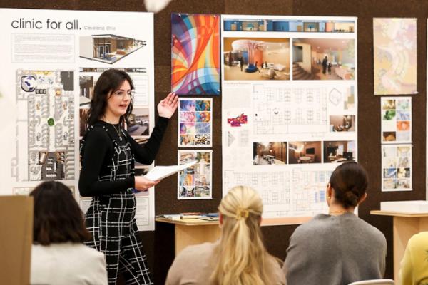 A student presents an interior design project during a review session. The student stands in front of display boards showing floor plans, renderings and color studies while speaking to a seated audience. The presentation boards feature healthcare-focused design concepts, including a project titled “clinic for all” and images of interior spaces.