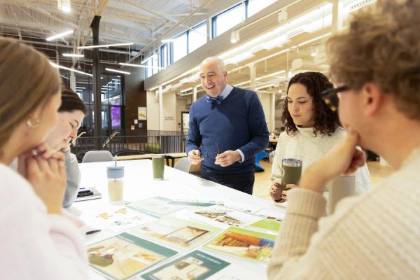 Students and an instructor gather around a table in a bright design studio, reviewing printed project materials and layouts. The group stands and sits around the table with notebooks and reusable drink cups nearby, while exposed beams, large windows and collaborative workspaces are visible in the background.