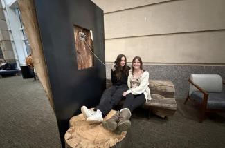 Two smiling people sit on a wooden bench in a spacious room, casually leaning against a vertical wooden art piece. The atmosphere is relaxed and informal.