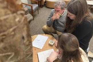 Three people focused on a brainstorming session at a round wooden table. Notes and measuring tape are present, creating a collaborative atmosphere.