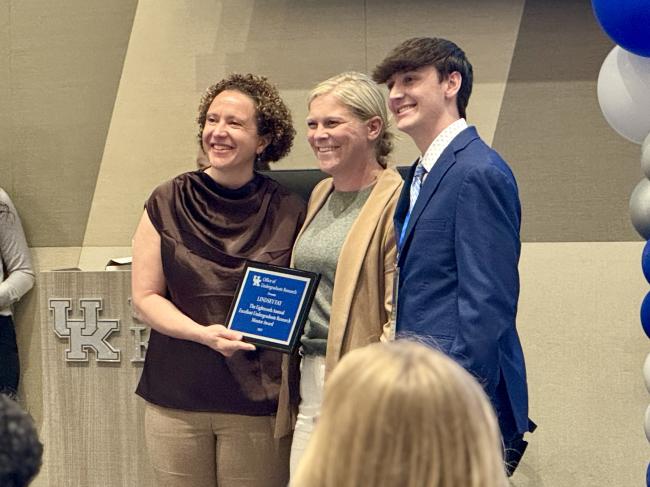 UK's Acting Vice President for Research Ilhem Messaoudi (left) and student nominator Brayden Nichols (right) present Lindsey Fay (center) with a commemorative plaque recognizing her as a 2025 Undergraduate Mentor of the Year..