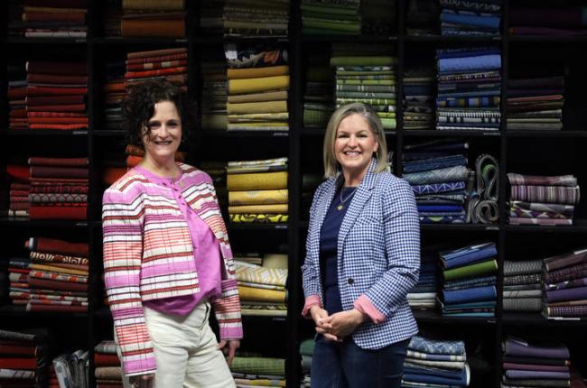 Two women stand smiling in front of shelves filled with colorful fabric rolls. Both wear patterned jackets, creating a vibrant and cheerful atmosphere.
