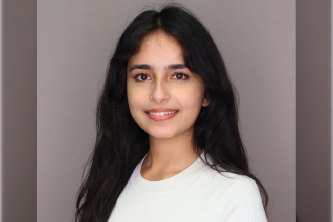 A woman with long dark hair and a subtle smile wears a white top against a gray background. She appears relaxed and approachable, conveying warmth.