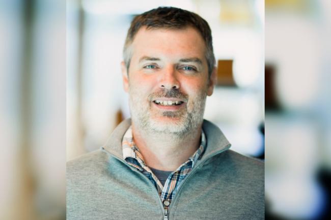 A man with short brown hair and a gray beard is wearing a gray zip-up sweater over a plaid shirt, smiling at the camera with a blurred indoor background.