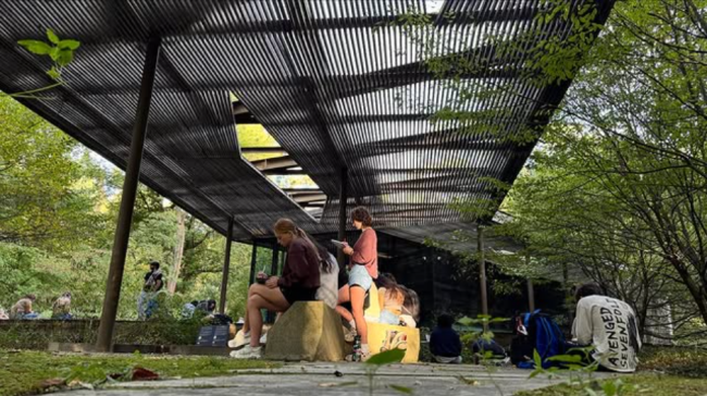 People gather beneath a shaded metal pergola in a leafy park, sitting on rocks and benches with backpacks, creating a relaxed outdoor mood.